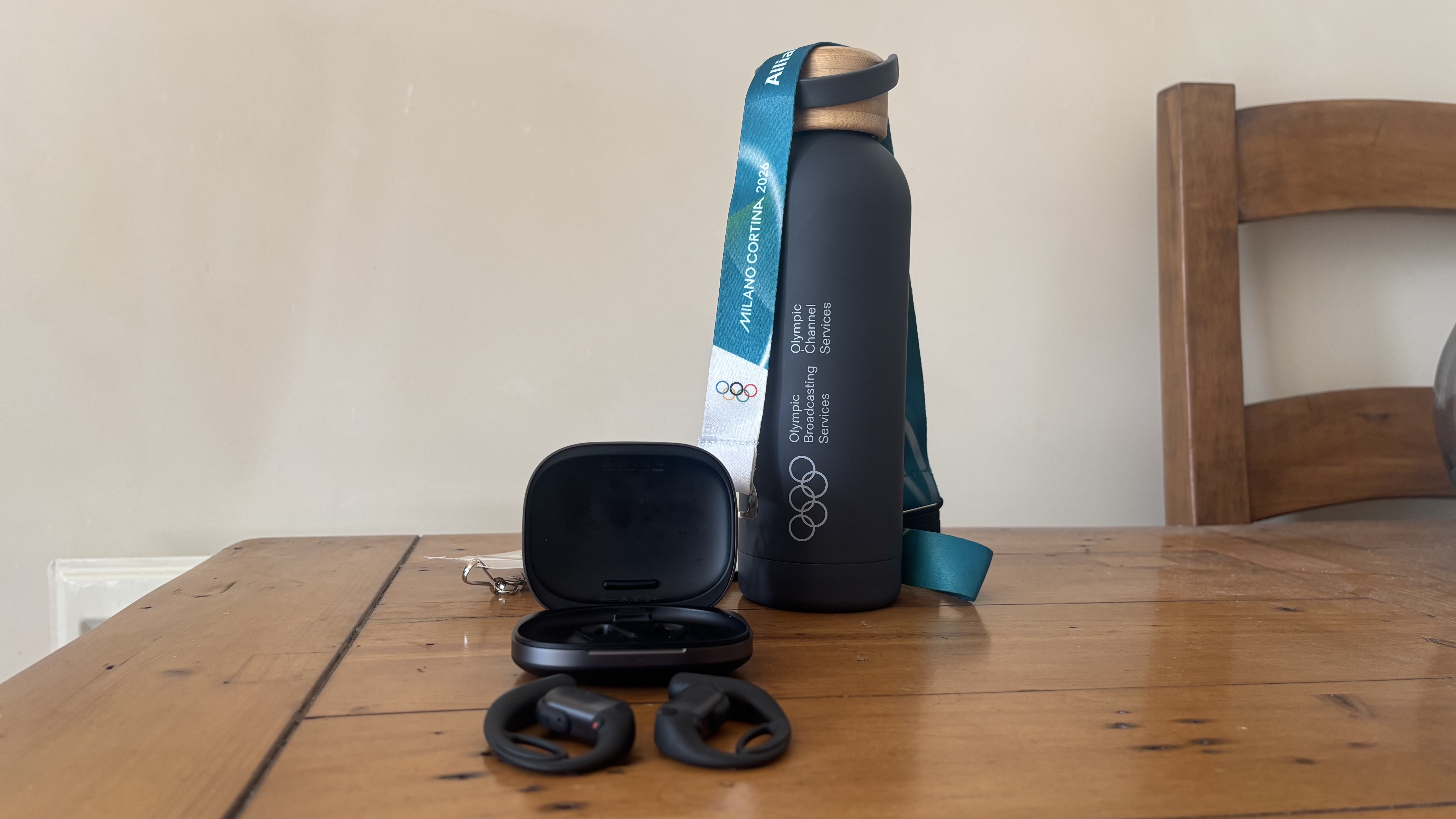 Shokz OpenFit Pro open earbuds on a wooden surface, in front of their carry case, and an Olympics water bottle and lanyard.