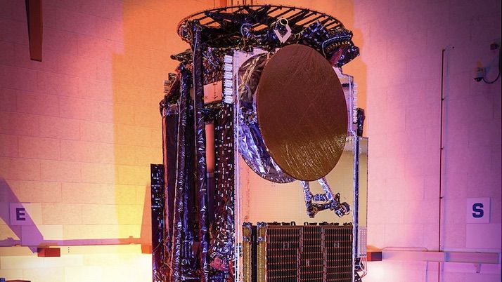 A towering satellite stands in a clean room facility, around hued lighting of oranges and pinks. 