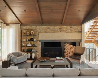 neutral living room with wooden ceiling, exposed stone wall with fireplace, neutral armchairs and sofas, coffee table and bookcase
