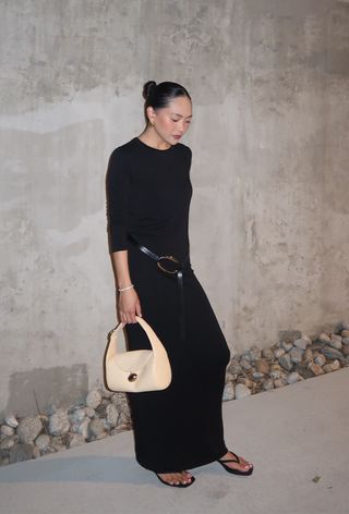 A woman wearing a black maxi dress, a black belt with gold hardware, a cream shoulder bag, and thong sandals.