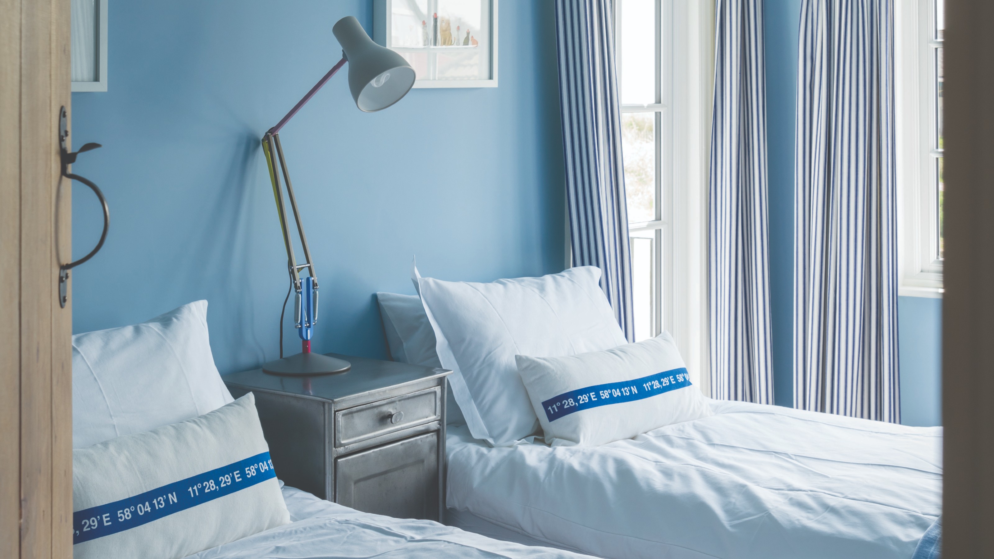 A light blue-painted twin bedroom in Farrow &amp; Ball's Lulworth Blue with striped white and blue curtains