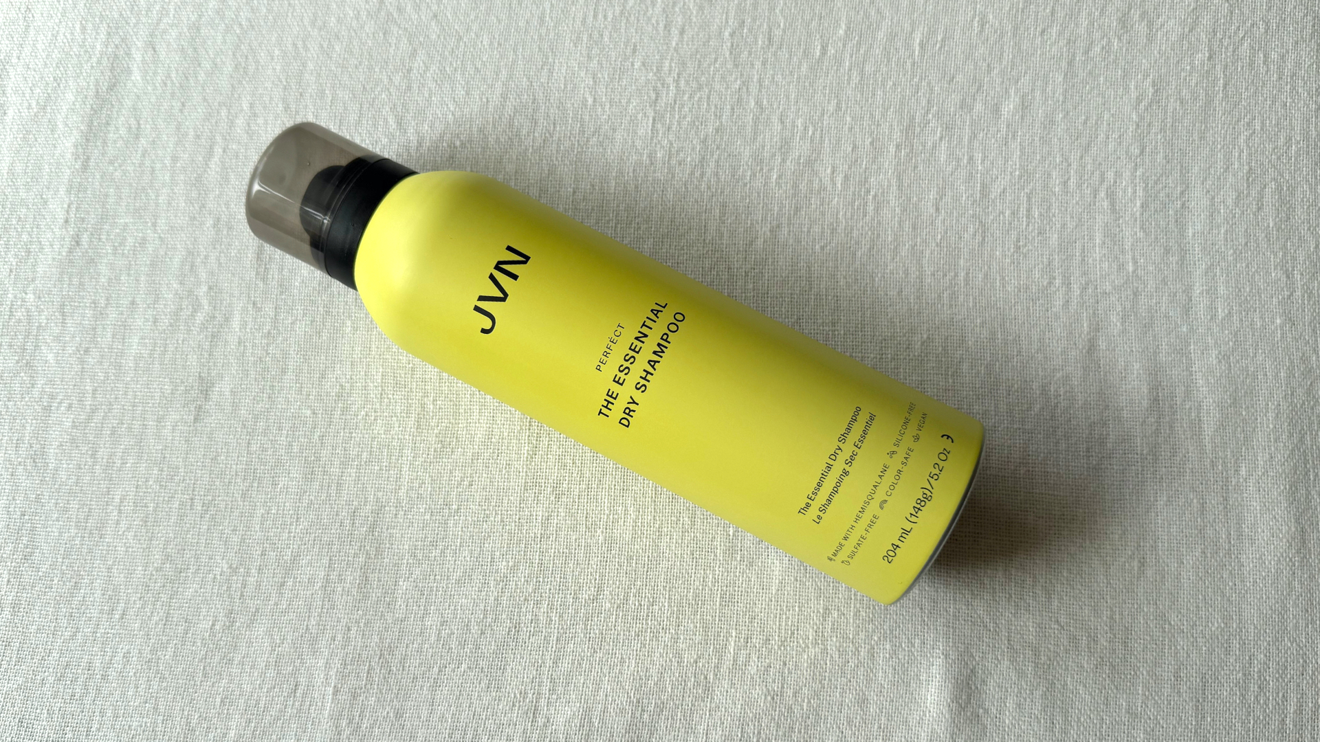 A bird's eye view of a can of JVN Hair The Essential Dry Shampoo laying at a 45 degree angle against an off-white linen background