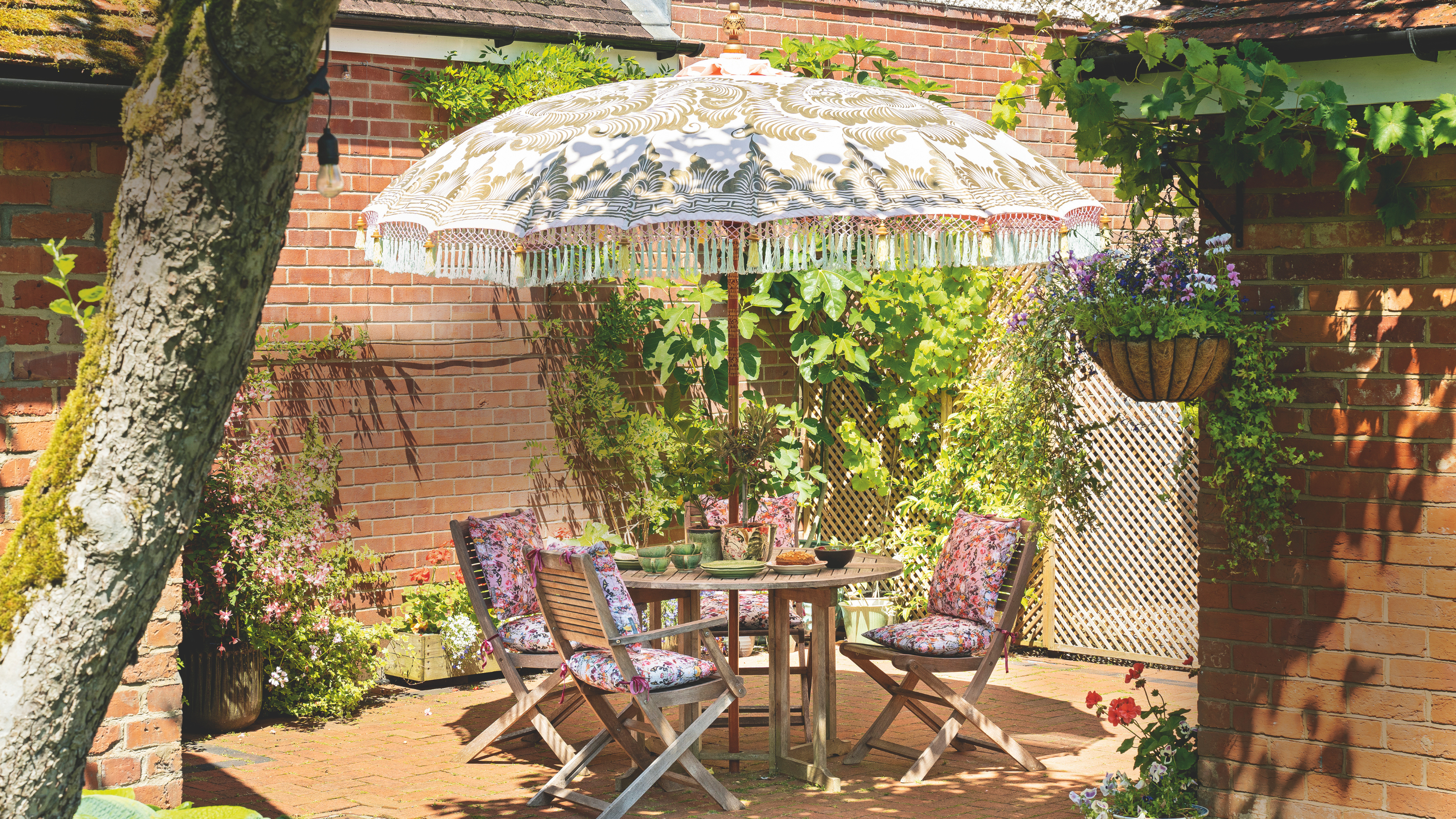 Patterned parasol and garden furnture in a courtyard with festoon lights and potted plants.