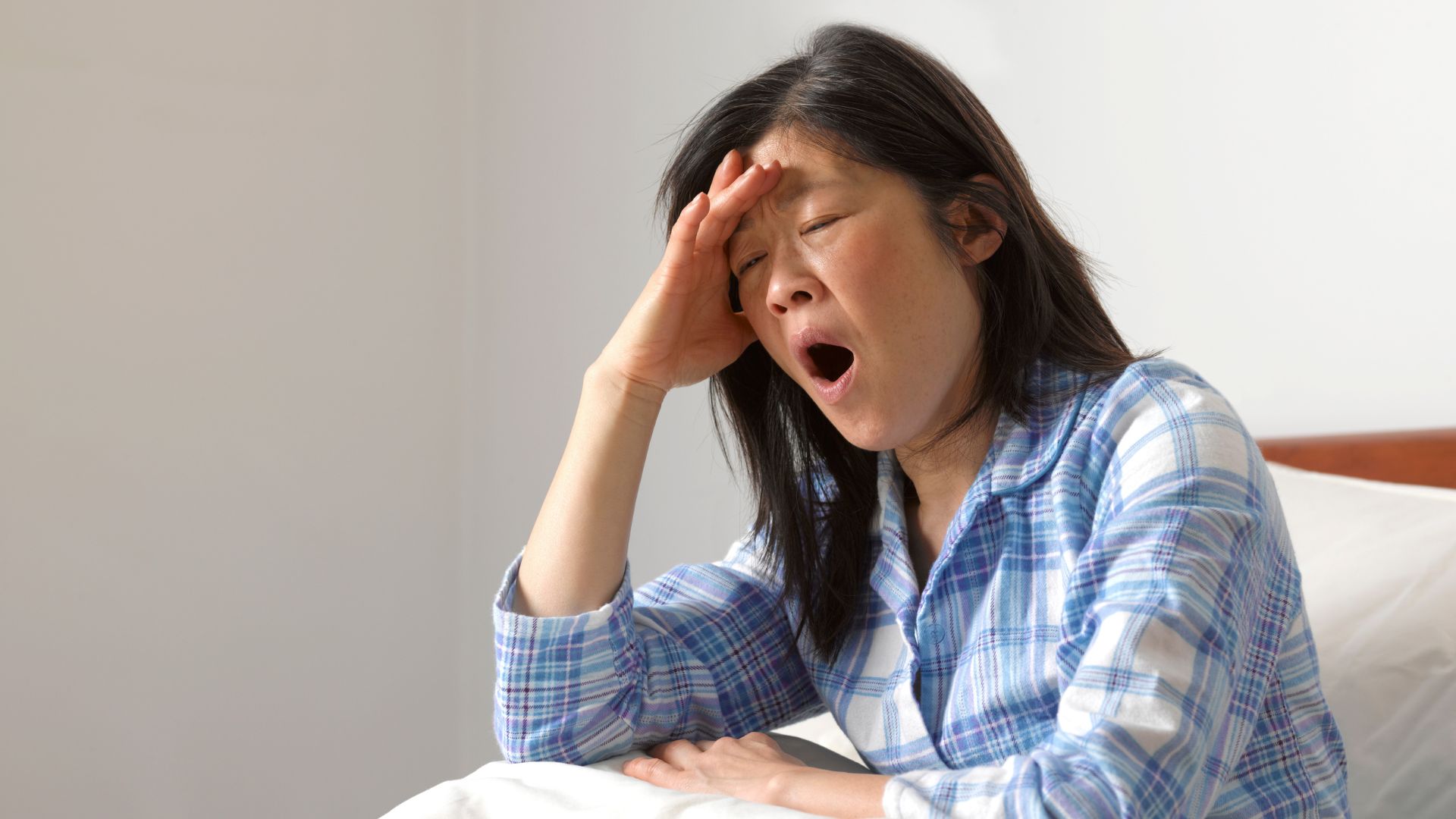 A woman in a blue and white checked pajama shirt sits in bed yawning.