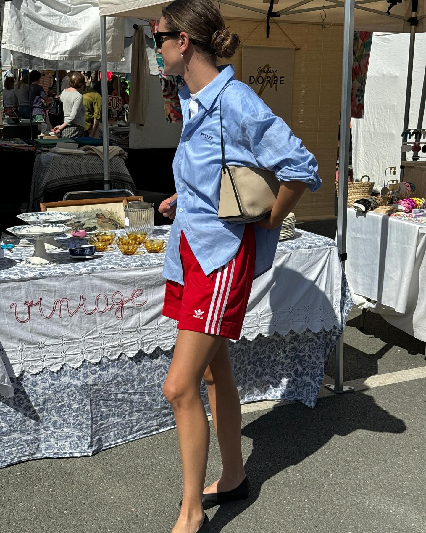 French fashion influencer Anne-Laure Mais poses at a farmer's market wearing a low bun, blue button-down shirt, tan shoulder bag, red striped Adidas track shorts, and black ballet flats