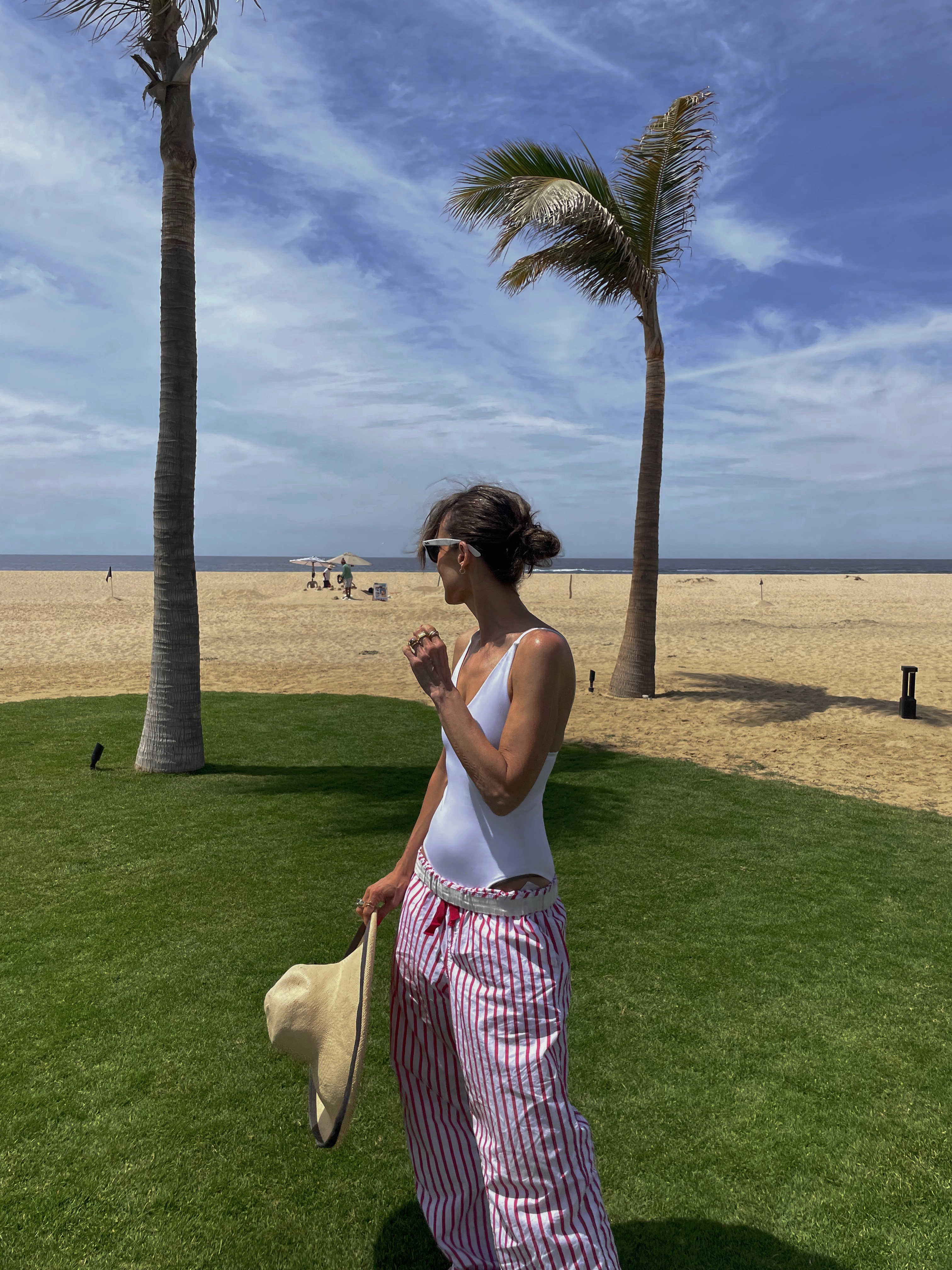 Larissa Mills in red and white striped pajama pants and a white bathing suit on the beach in front of a palm tree