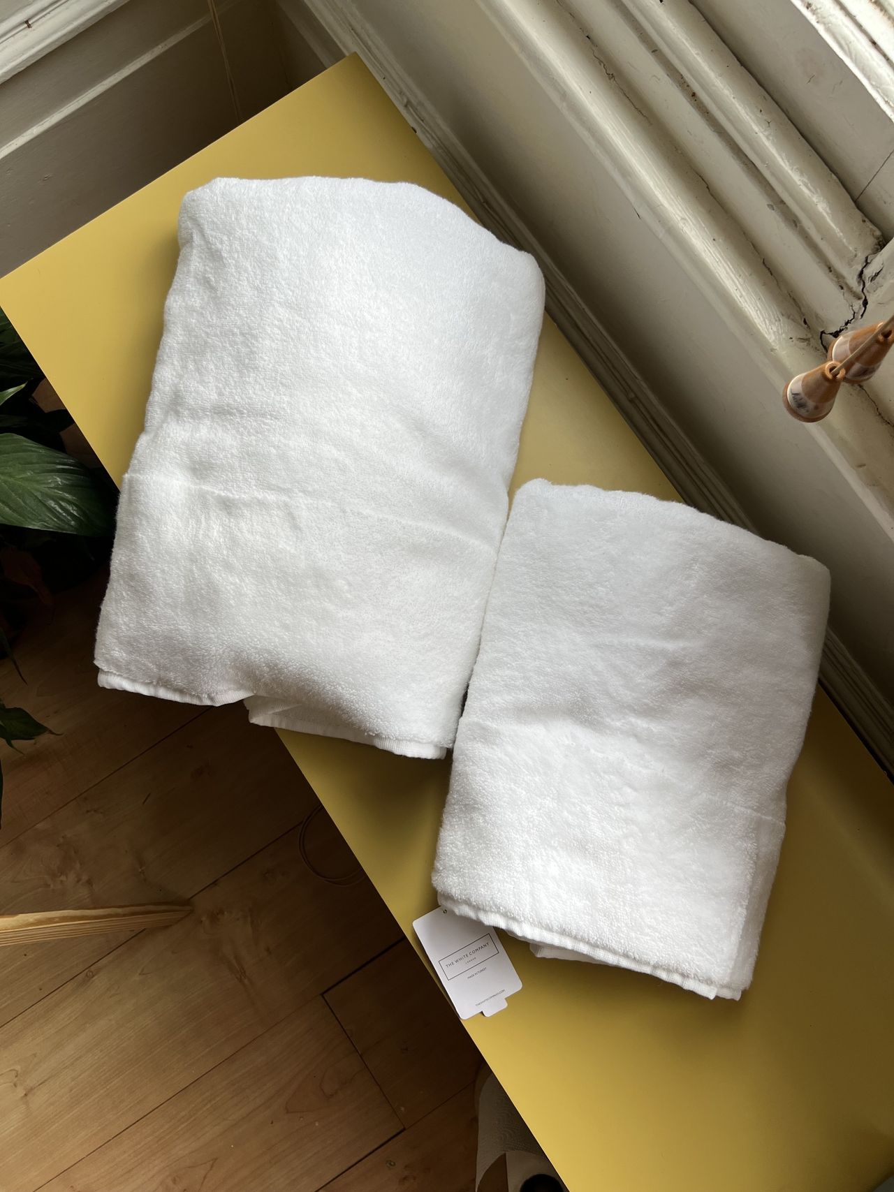 Image of two different-sized white towels folded on a long yellow console by a window.