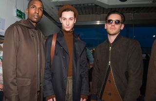 Three male models wearing looks from Oliver Spencer's collection. One model is wearing a dark coloured shirt and brown coat. Next to him is a model wearing a brown hat, brown jumper, multicoloured striped trousers, blue coat and brown backpack. And the third model is wearing sunglasses, a brown cardigan and brown partially zipped jacket on top