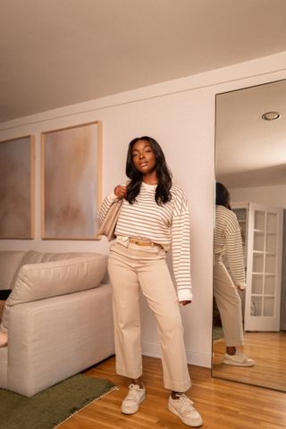 Woman standing in apartment wearing striped crewneck sweatshirt, khaki cropped pants, brown belt, white and tan sneakers