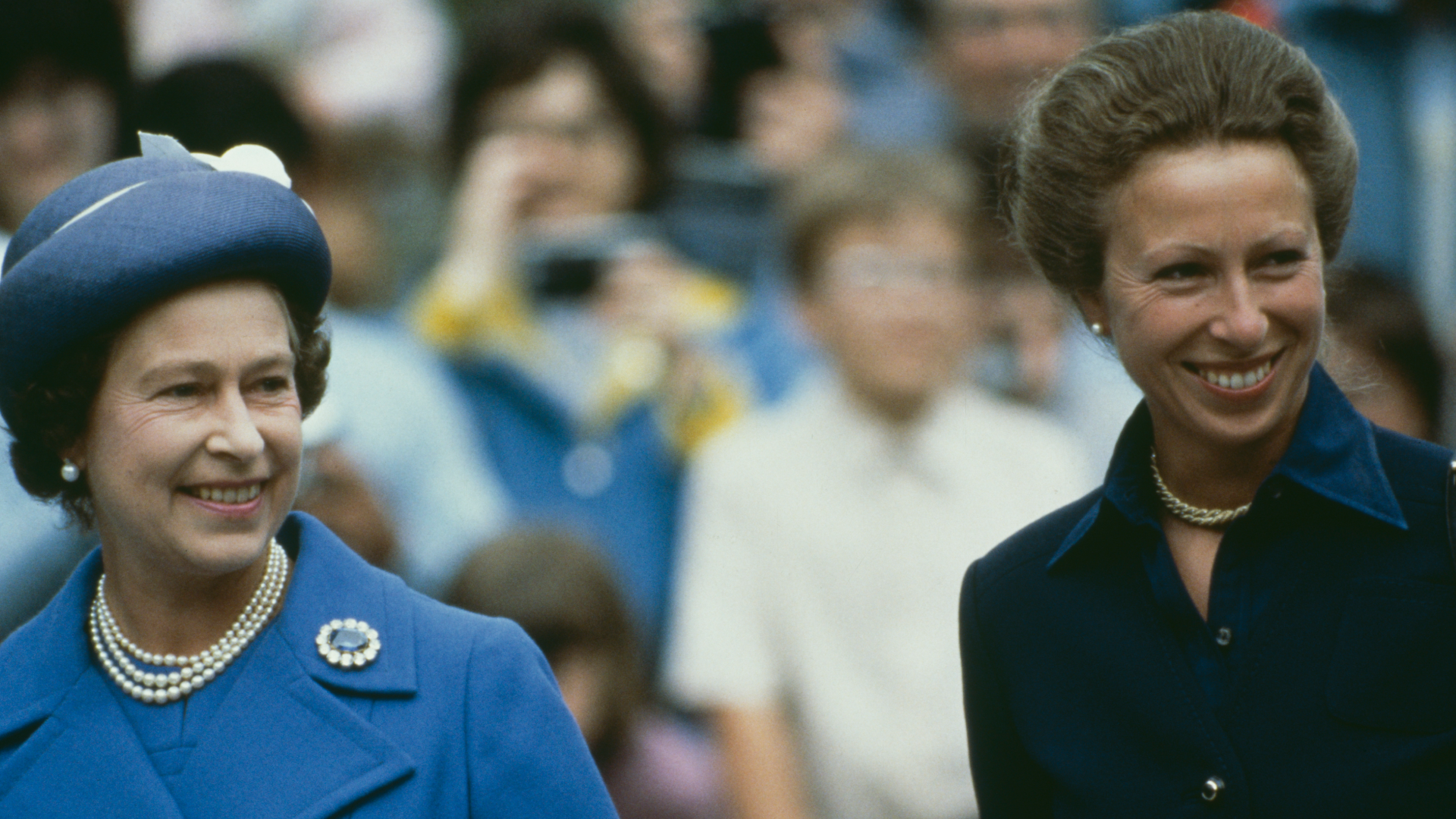 Queen Elizabeth wearing a blue coat and hat standing next to Princess Anne