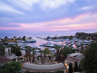 Sani Resort's private Marina at sunset. Yachts and boats line the water, the restaurants and shops are lit up and the sky is pale pink, yellow and violet