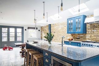 Industrial style kitchen extension in london, with blue units, and a large island with wooden stools, an exposed brick wall and blue doors onto the outside space