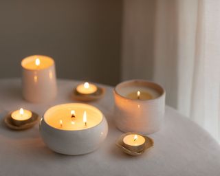 A collection of white candles burning on a white table with a grey wall and white curtain in the background