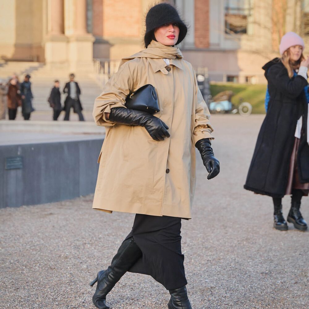 A woman walks with a trench coat, leather gloves, black leather clutch, textured hair bucket hat, and black boots. 