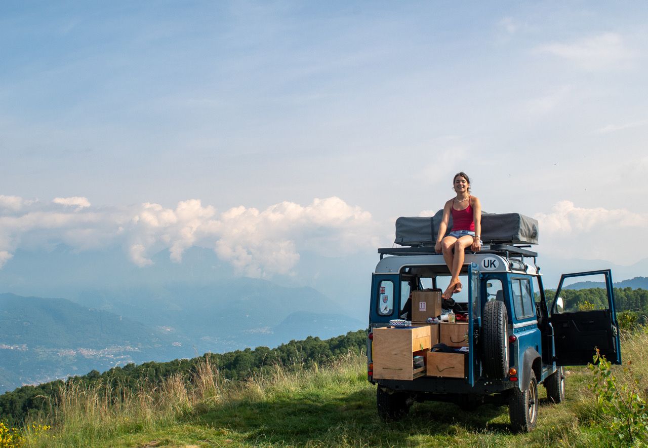 Tati Reed sits on top of Blue Tit, the Land Rover Defender, on a scenic European hill