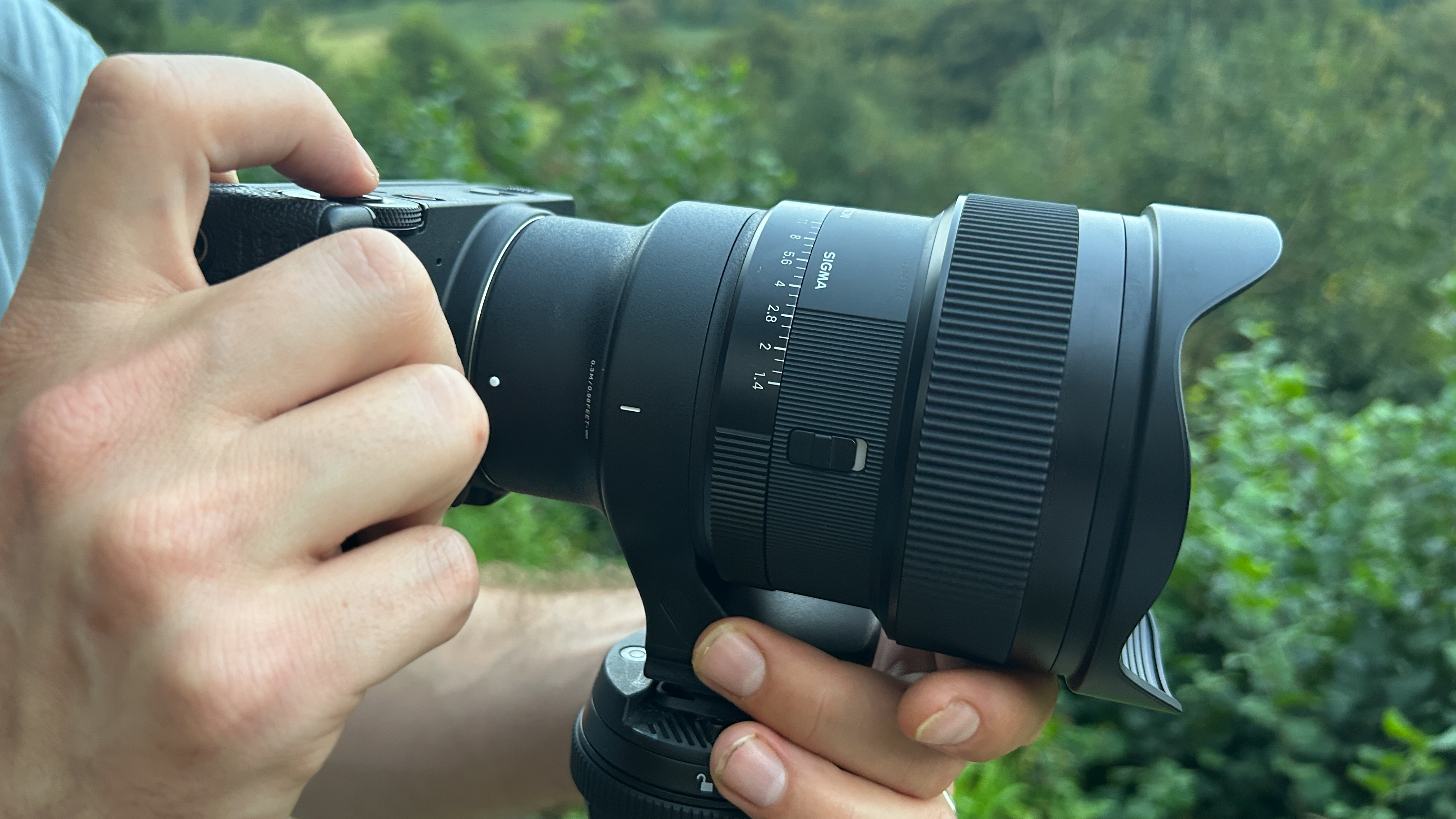 A black camera lens with two ridged adjustment rings is mounted to an out-of-frame tripod and attached to a rectangular black camera. Hands support the item at the mount base of the tripod and on the camera. An index finger rests on the camera trigger button.