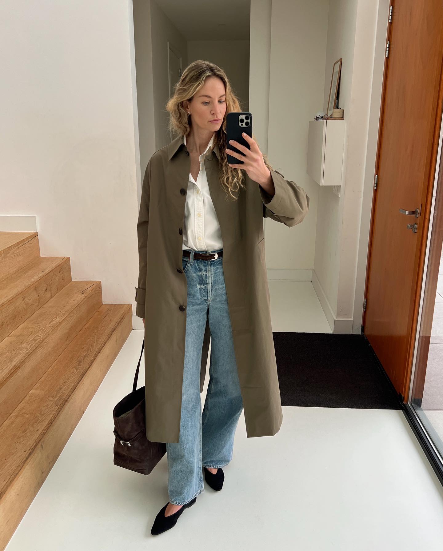 a stylish woman poses for a mirror selfie wearing a green trench coat, white button-down shirt, skinny belt, straight-leg jeans, The Row Margaux bag, and pointed-toe flats