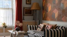 Living room with pale blue walls, striped sofa, red curtains, a glass and stone coffee table, a large painting above the sofa in orange and red of houses, and a rattan floor lamp in the corner