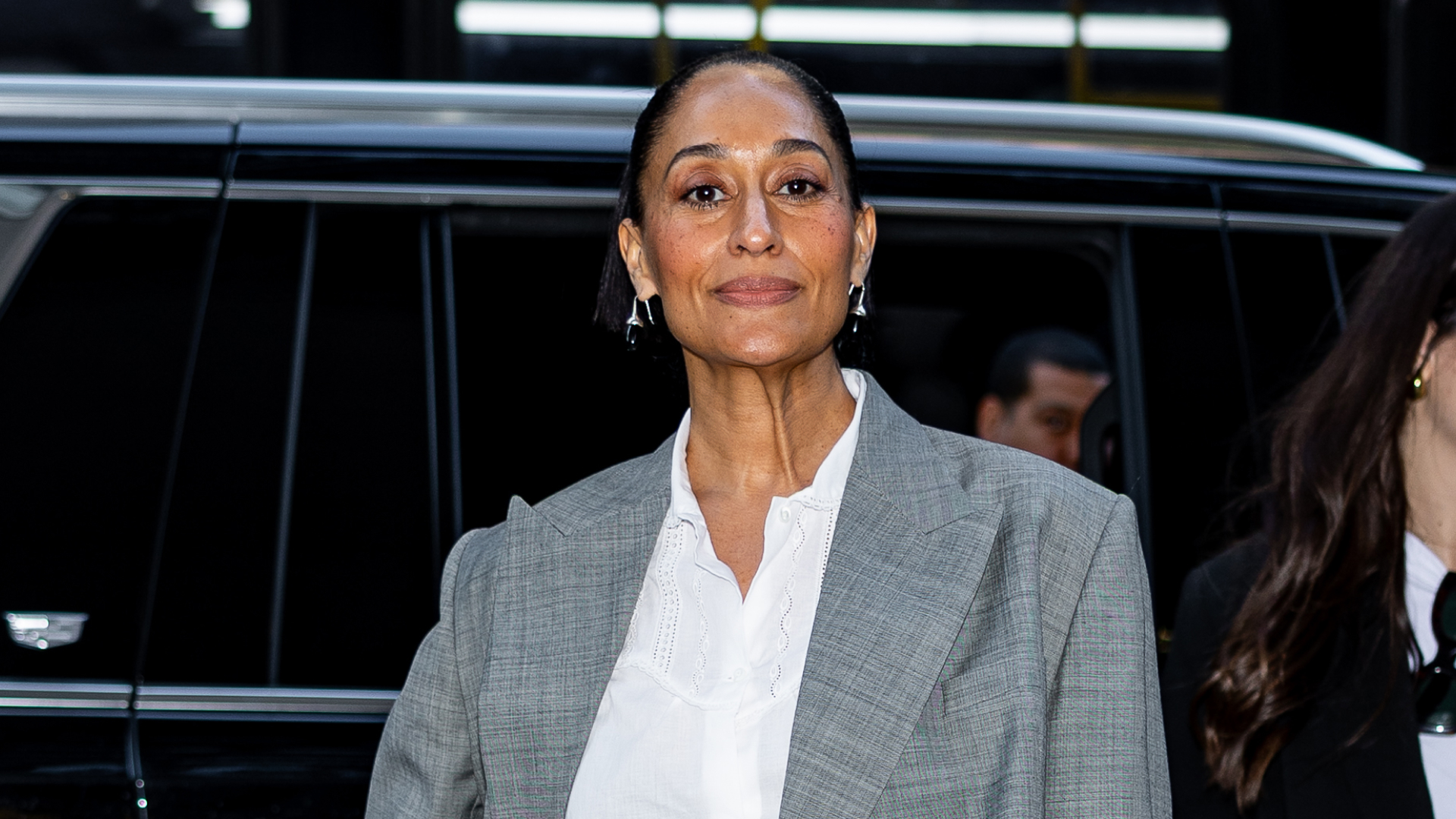 Tracee Ellis Ross wearing a white button up under a grey blazer. She is wearing a slicked back bun and silver hoop earrings.