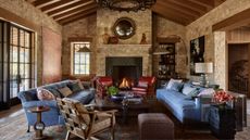 a stone barn style vaulted living room with large fireplace with a lit fire, blue sofas facing inwards with leather chairs and cozy interiors
