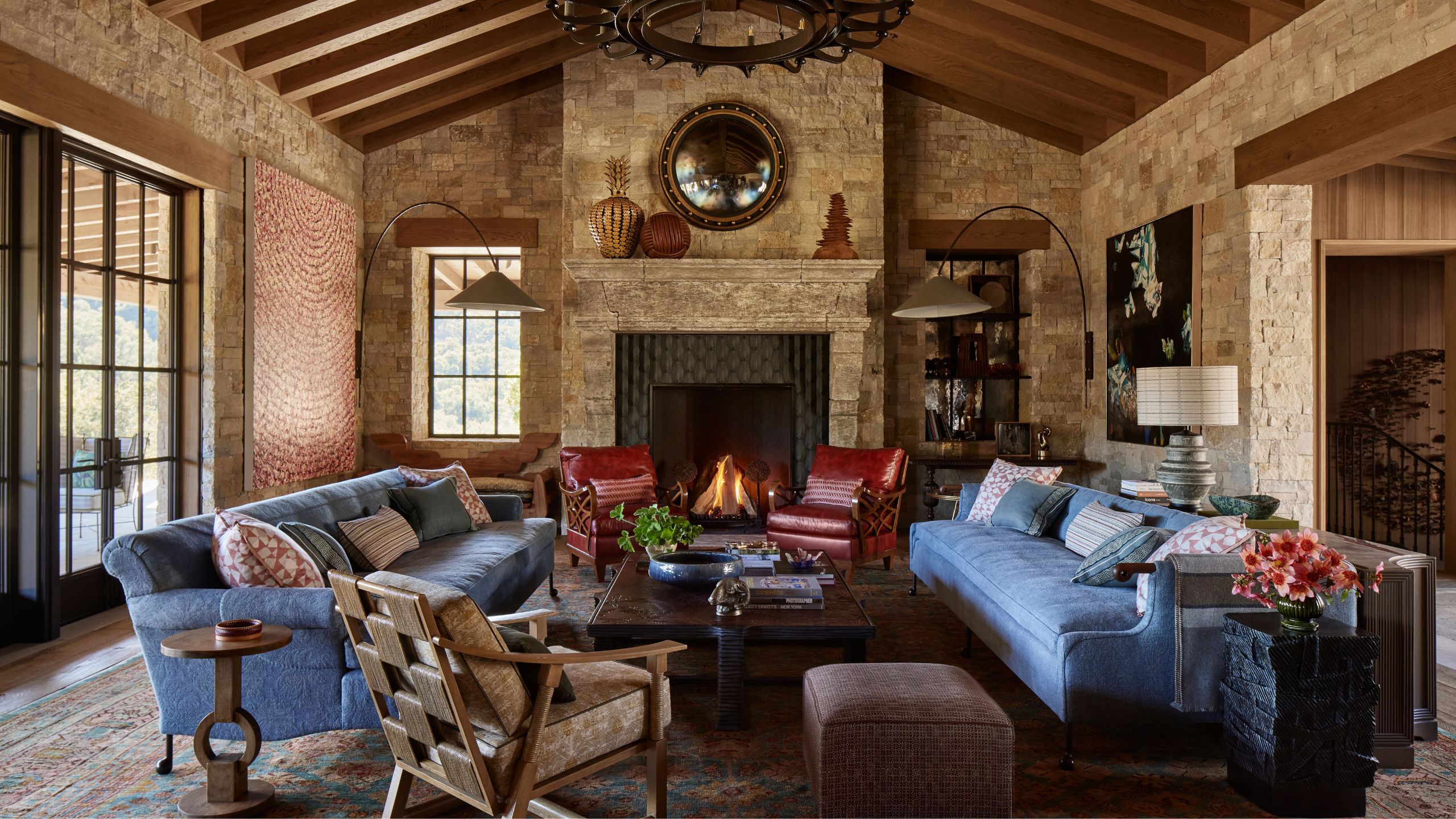 a stone barn style vaulted living room with large fireplace with a lit fire, blue sofas facing inwards with leather chairs and cozy interiors