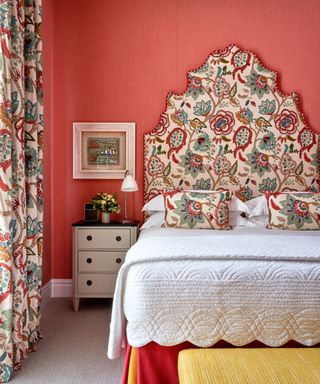 red bedroom with stylised floral headboard and matching drapes, white bedding, yellow ottoman, artwork