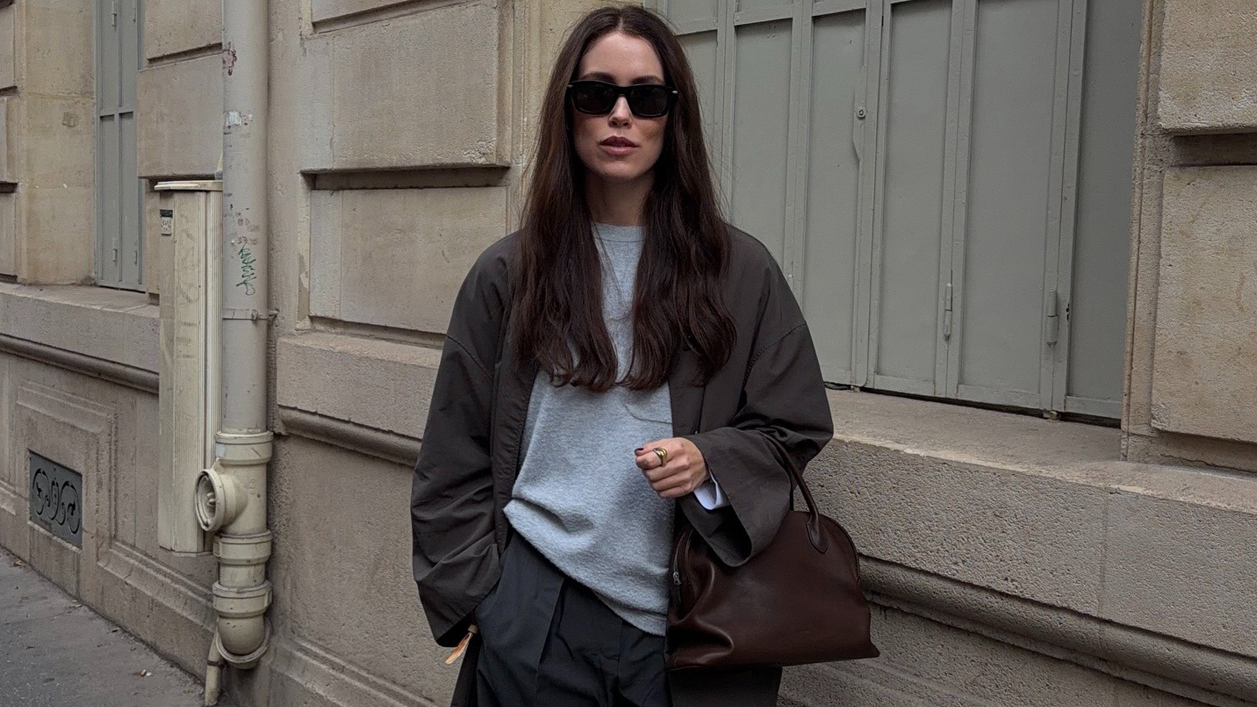 Annabel wears a brown long coat, gray t-shirt, gray trousers, and a brown the row bag.