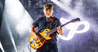 Joey Santiago wears a Castro cap and plays a Bigsby-equipped Goltdtop, onstage with the Pixies. The Pixies&#039; logo is illuminated behind him.