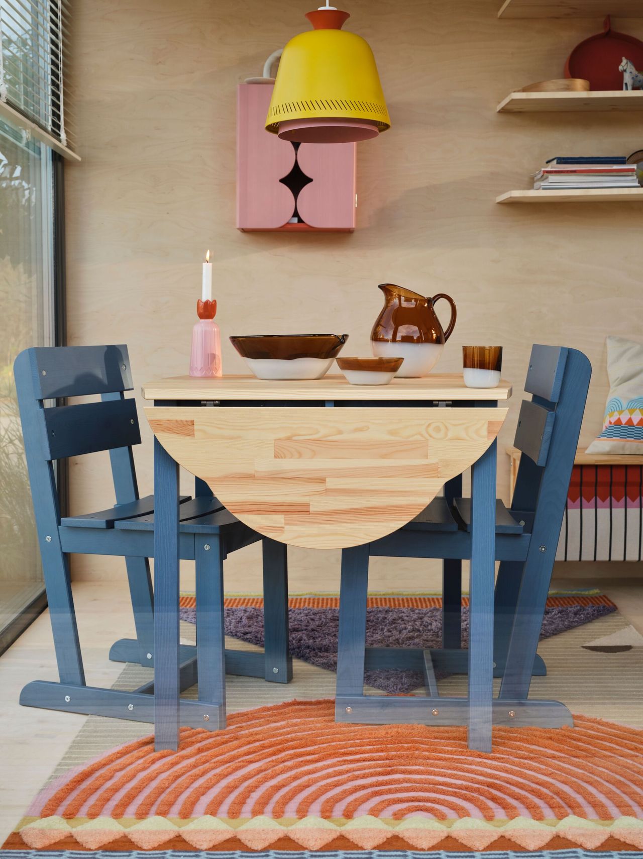 Image of a wooden table with blue legs and matching blue dining chairs. There is amber glass serveware on the table and a pink and orange candle holder. There is a pink and orange woven patterned rug underneath.