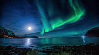 green northern lights shimmer over a body of water with the bright moon above