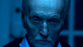 Tobin Bell grimaces while sitting in a blue lit room in Saw X.