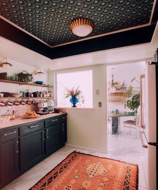White kitchen with black, scalloped wallpaper design on ceiling, gold ceiling light, pink patterned rug on floor, black kitchen cabinets, looking out onto dining room space