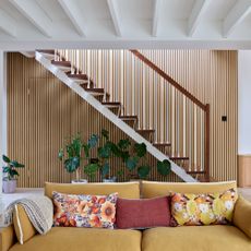 An open-plan living room with a mustard yellow sofa with a staircase and a vertically panelled wall behind it