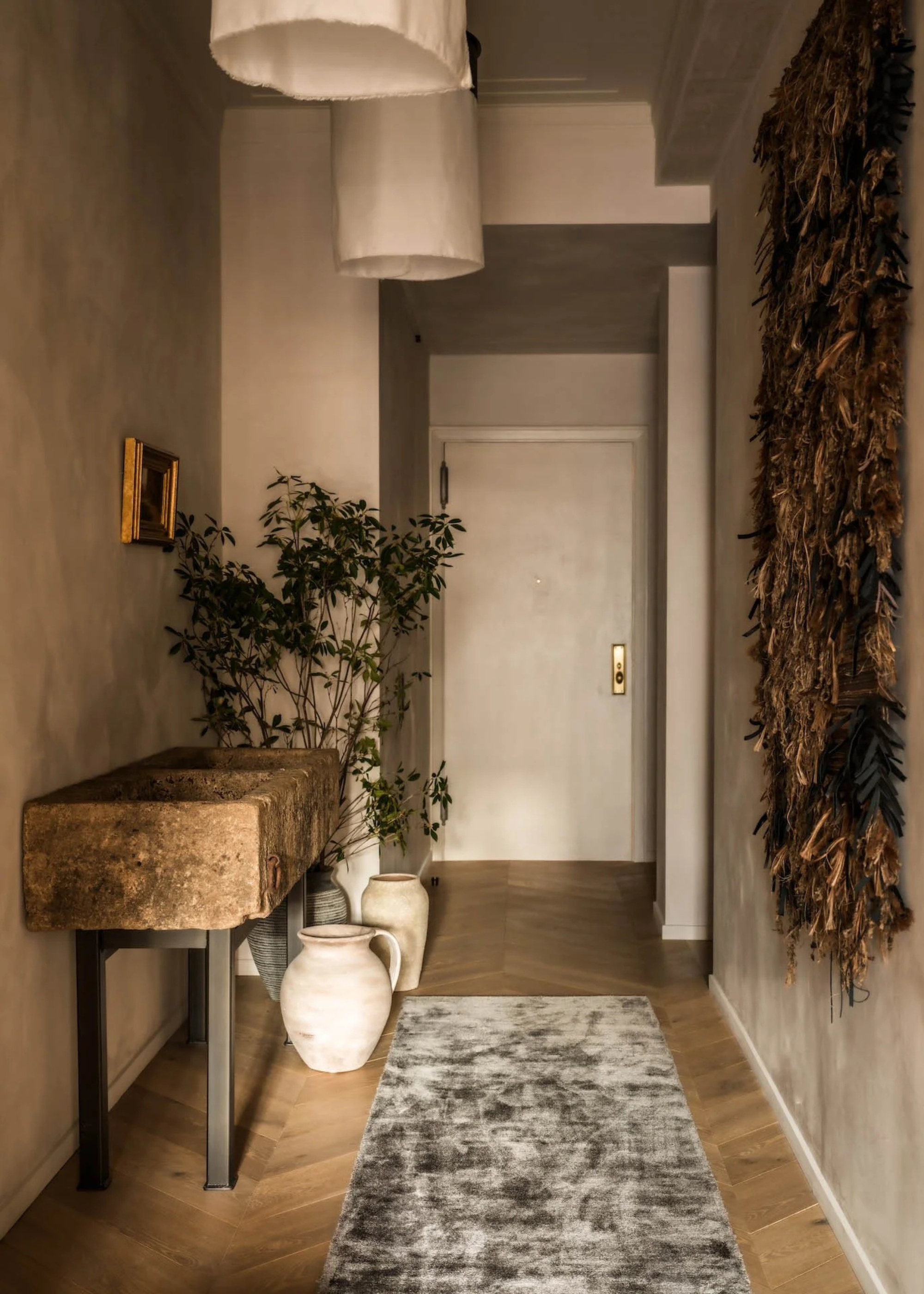 A limewashed entryway with a stone side table, vases of stems, a velvet runner on the herringbone wood floor and a feathered tapestry on the wall