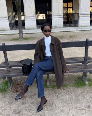 Woman wearing a white thin turtleneck with jeans and a long brown coat.