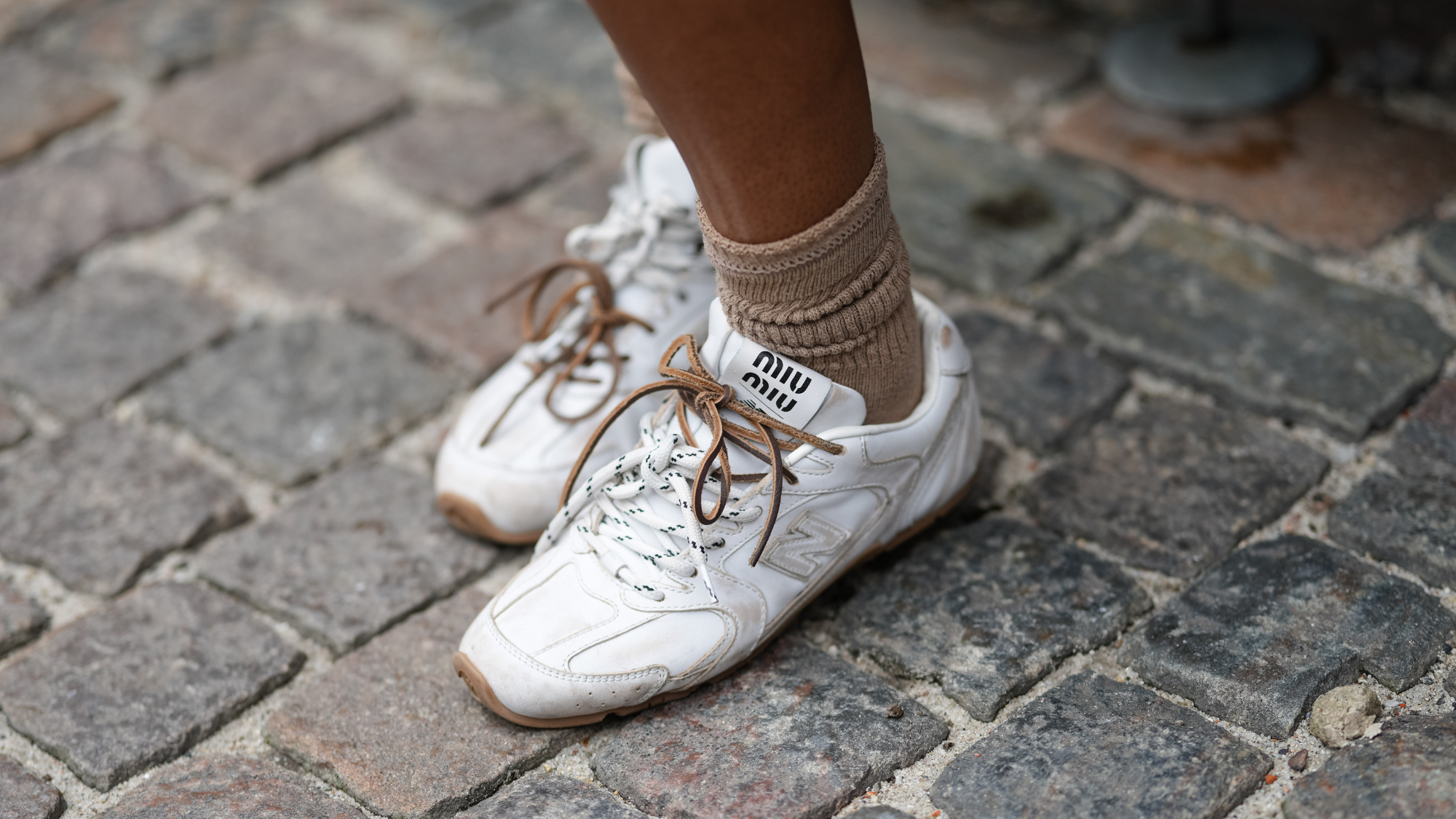 A guest wears white Miu Miu sneakers with a textured leather finish, featuring a prominent logo on the tongue. The sneakers have brown laces and a light brown sole. Paired with the shoes are ribbed socks in a beige tone, outside Forza Collective, during Copenhagen Fashion Week