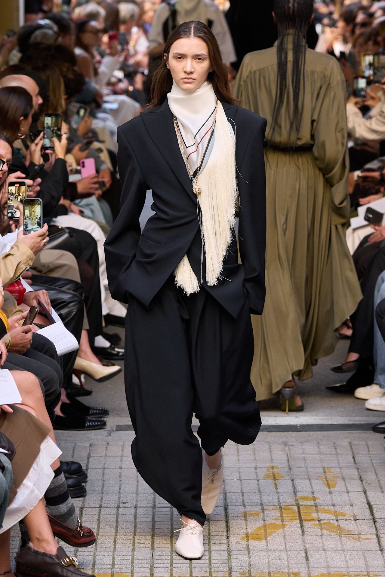 Layering staples for spring 2026 is shown in a photo from Calcaterra's runway show, with a model walking while wearing a black blazer styled with a white fringe scarf around the neck, black trousers, and white derby shoes