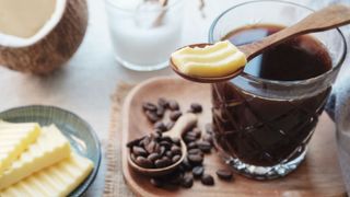 The components of bulletproof coffee scattered around a cup of black coffee: a teaspoon of butter, half a coconut, coffee beans and a cup