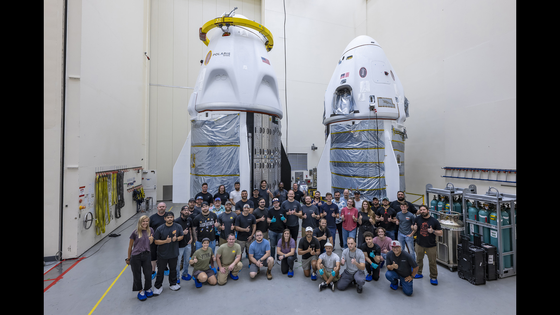 In a large room with white walls you can see two white space capsules in front of which there is a large crowd of people