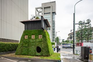 Displayed on a side-walk and on the corner of a junction, a c.10m tall narrow house with a large base than top. The first half is covered in grass with three square windows on the left side and front. There is a circular hole at the bottom of the grass where a door would be. The top third of the art piece if a wooden structure with a roof.