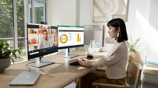 De bedste PC-skærme: En kvinde sidder og arbejder med to skærme på et lyst hjemmekontor foran store vinduer