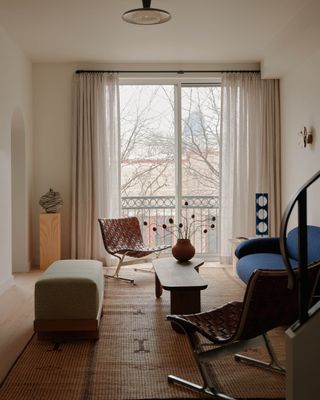 small living room with windows opening out to balcony, plinth in corner, long ottoman, two woven armchair, timber coffee table, and blue sofa