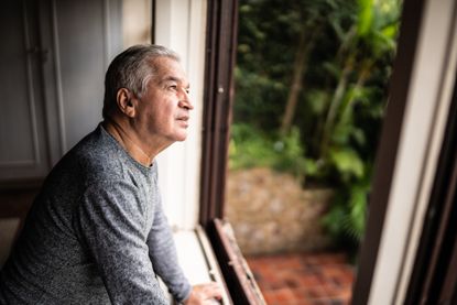 An older man is contemplating while looking through a window to the outdoors. 