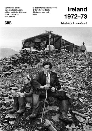 Publication cover with a black and white photograph of a man holding a stick and a young girl sat next to him amongst a pile of rubble. Behind them, there is a makeshift shelter and a woman with her back turned.