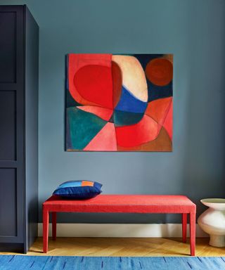 Blue painted hallway with wooden flooring, blue rug. Bright red bench, bold, colorful artwork mounted on wall above bench, painted dark blue cabinet in corner