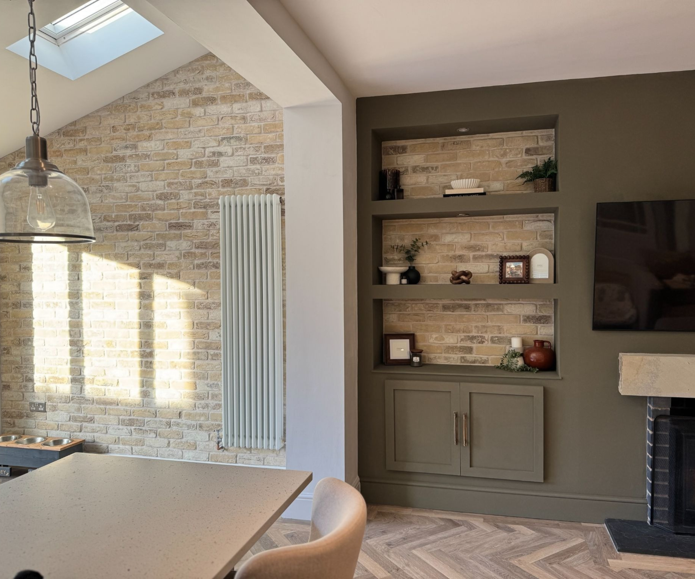 Open-plan kitchen-living space with exposed brick, built-in olive-green shelving, herringbone floor, and a fireplace.
