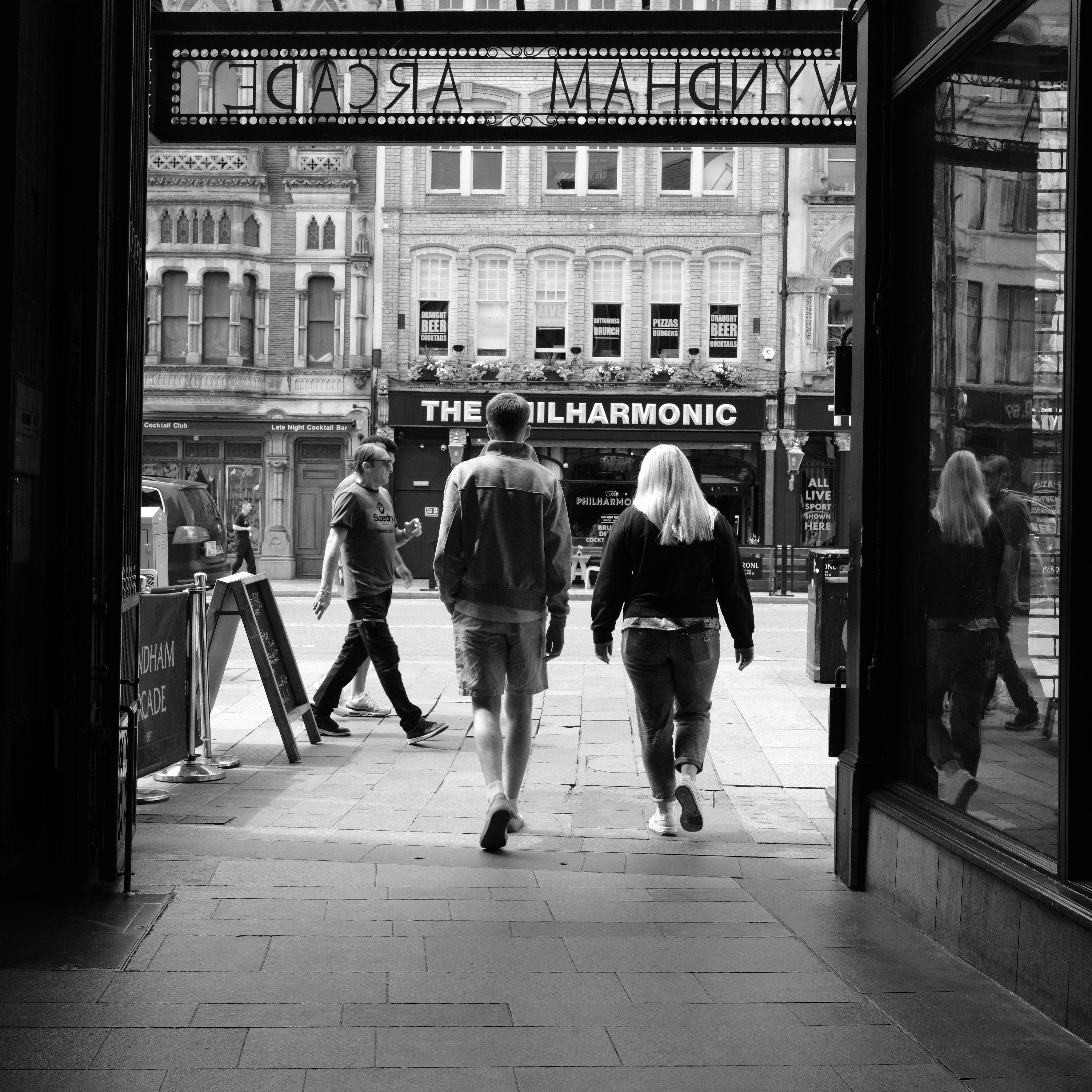 A black and white street scene of two people walking from a dark arcade into sunlight, taken on the Fujifilm X-E5.