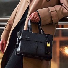 A person wearing a brown trench coat, holding a black tote bag from TUMI.