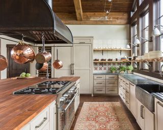 Large rustic kitchen, gray cabinetry, kitchen island with wooden countertop, pot rack hacking above, shelves decorated with kitchenware and glassware, rug on stone floor, wooden beamed ceiling