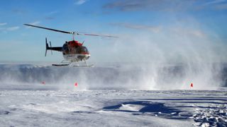 A red helicopter hovers over a large ice sheet, kicking up snow with its propellers